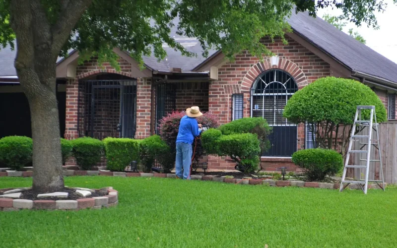 Landscaper trimming hedges in front of a brick house, showcasing well-maintained greenery and lawn, highlighting landscaping services and automation benefits.